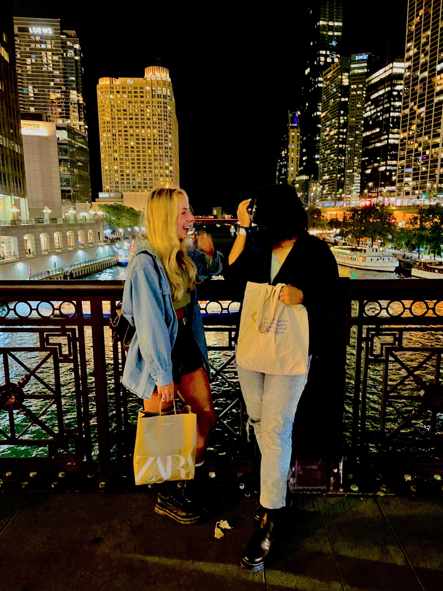 Zara and friend with shopping bag on bridge overlooking skyscrapers at night.