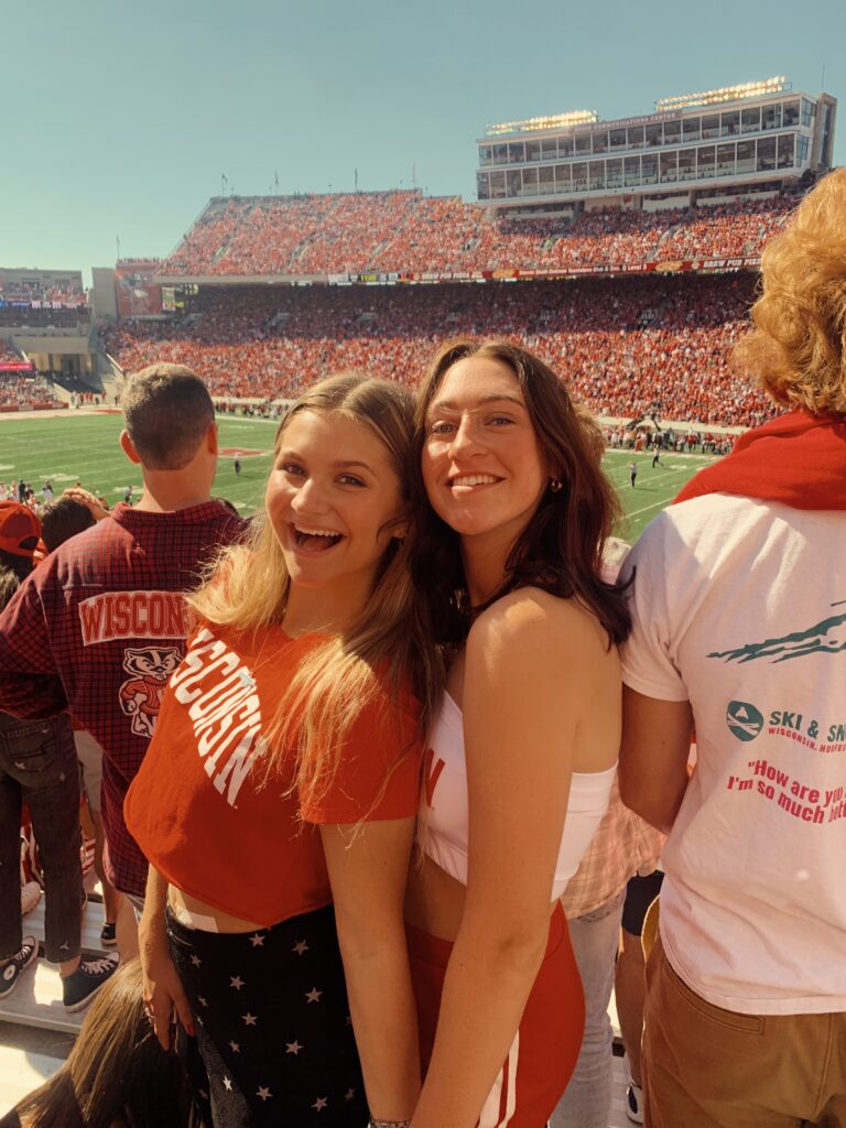 Two smiling girls wearing red and white at sunny football game with filled stadium seats in background