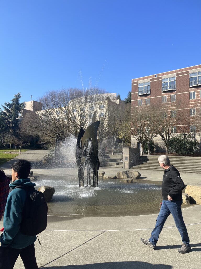 Students walking by abstract fountain in sunny quad with two academic buildings in background.