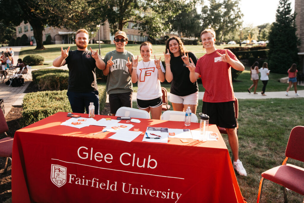 Five smiling students behind Glee Club information table with sunny campus in background