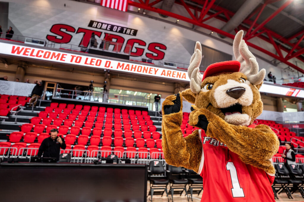 Stag mascot in arena in front of empty red seats and Home of the Stags signage