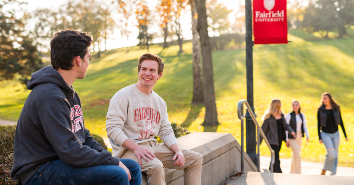 Smiling students in Fairfield sweatshirts talking while sitting on concrete wall on sunny fall day